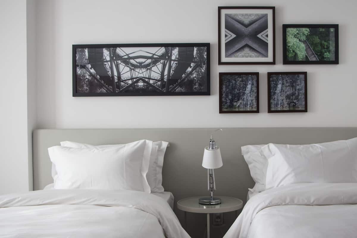 A hotel room with a bed, two pillows, a bedside table with a lamp, and four framed pictures on the wall.