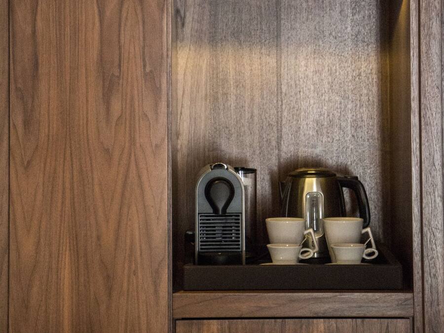 Un mueble de madera con dos estantes. El estante superior sostiene un jarrón blanco y un cuenco blanco. El estante inferior contiene una radio antigua, una cafetera y un juego de tazas y platillos blancos.