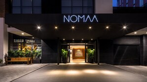 Entrada de hotel con un letrero de neón que dice "NOMAA", un banco de madera y plantas en macetas.