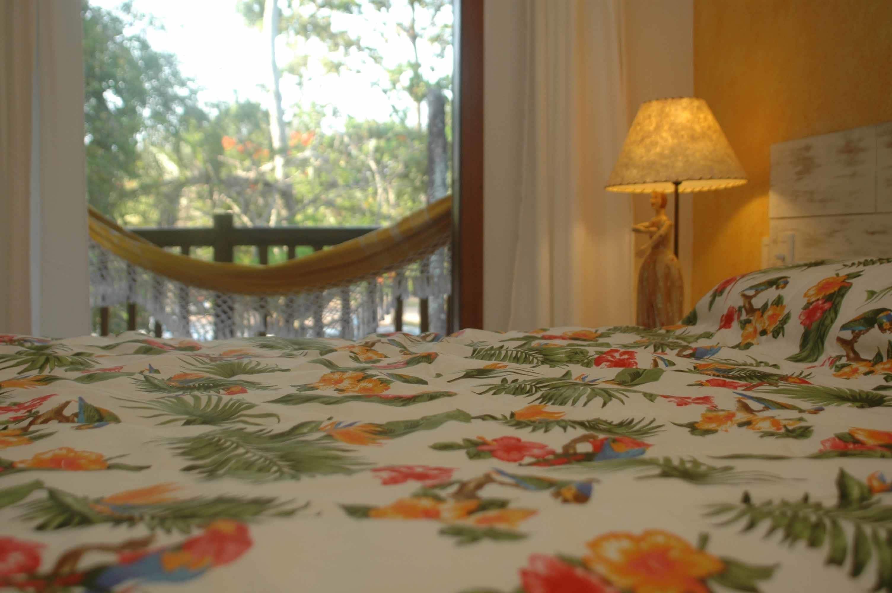 Uma cama com colcha florida, uma rede, um abajur e vista para árvores pela janela.