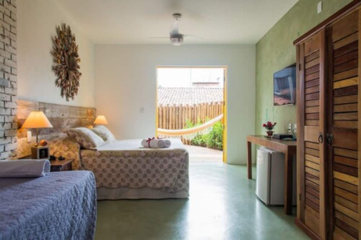 A bedroom with two beds, a small table with a lamp, a wooden door, and a view of the outside.