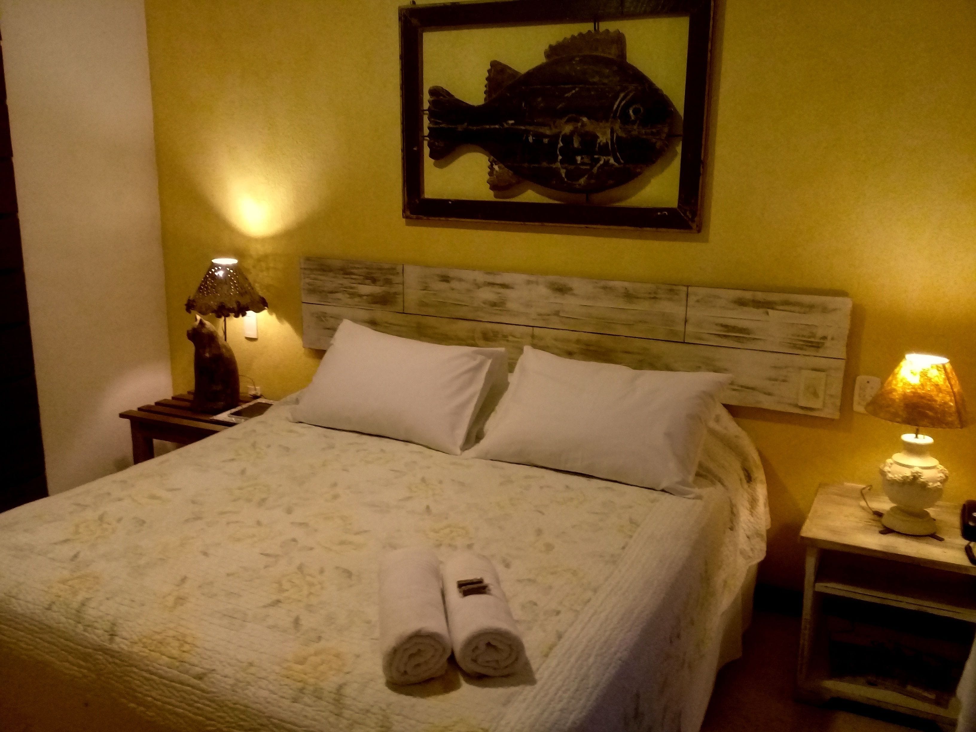 A hotel room with a bed, two pillows, a headboard, a nightstand with a lamp, a framed fish artwork on the wall, and rolled towels on the bed.