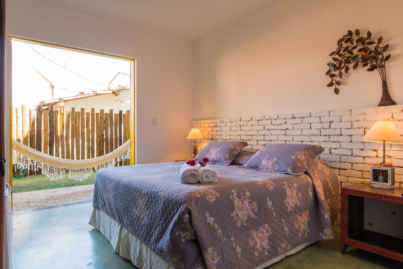 A bedroom with a bed, two bedside lamps, a hammock outside, and a brick wall.