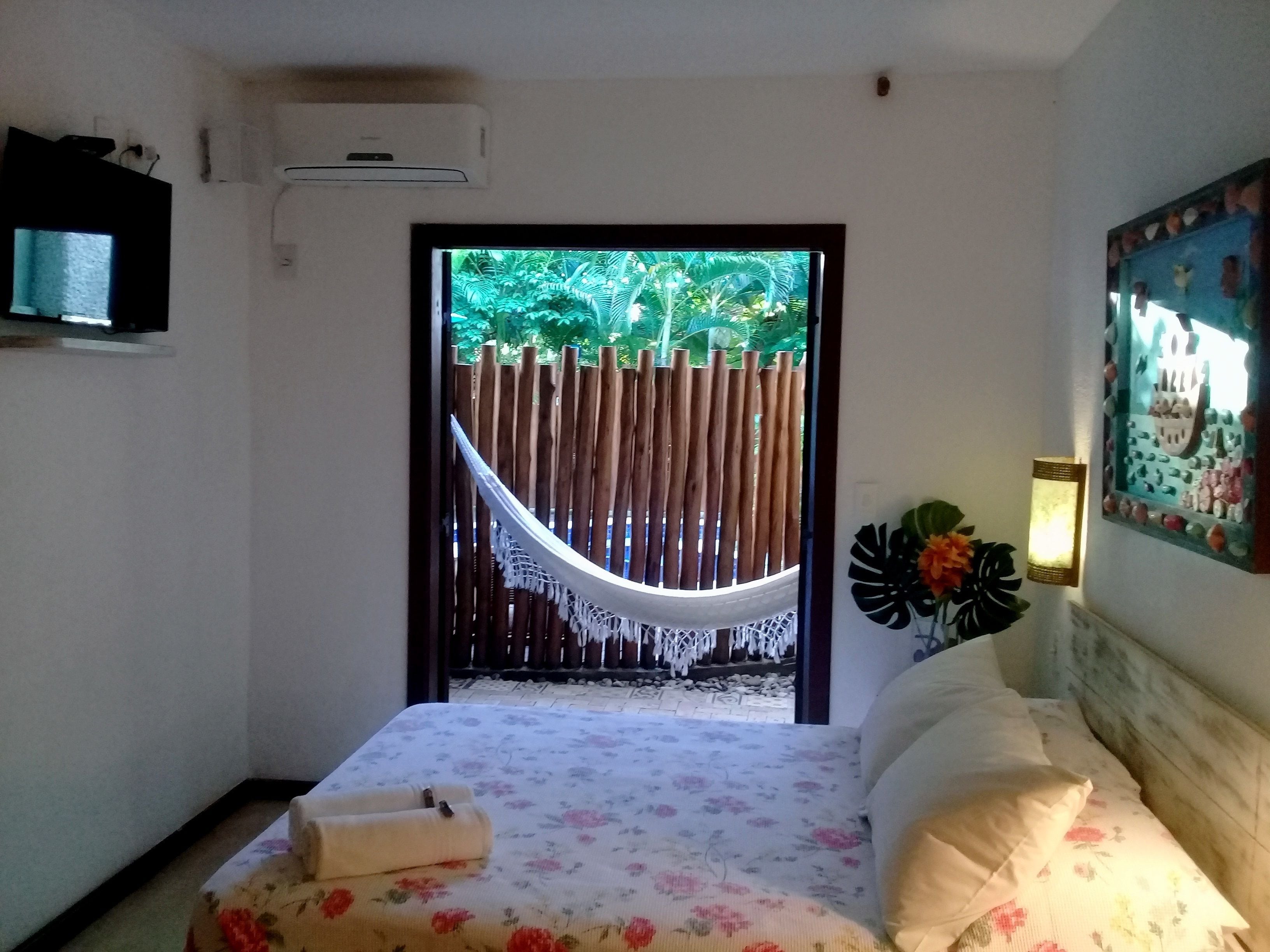 A bedroom with a bed, a TV, a hammock, and a view of greenery outside.