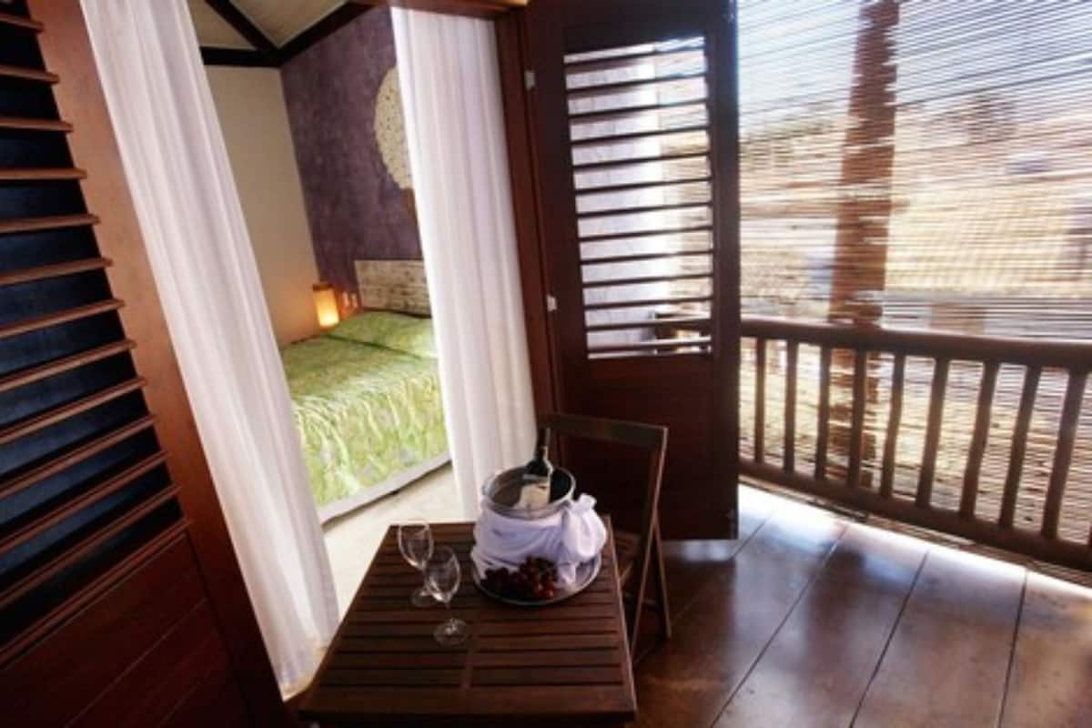 A wooden balcony with a table set for two, a bed with a green bedspread, and a view of the outdoors.