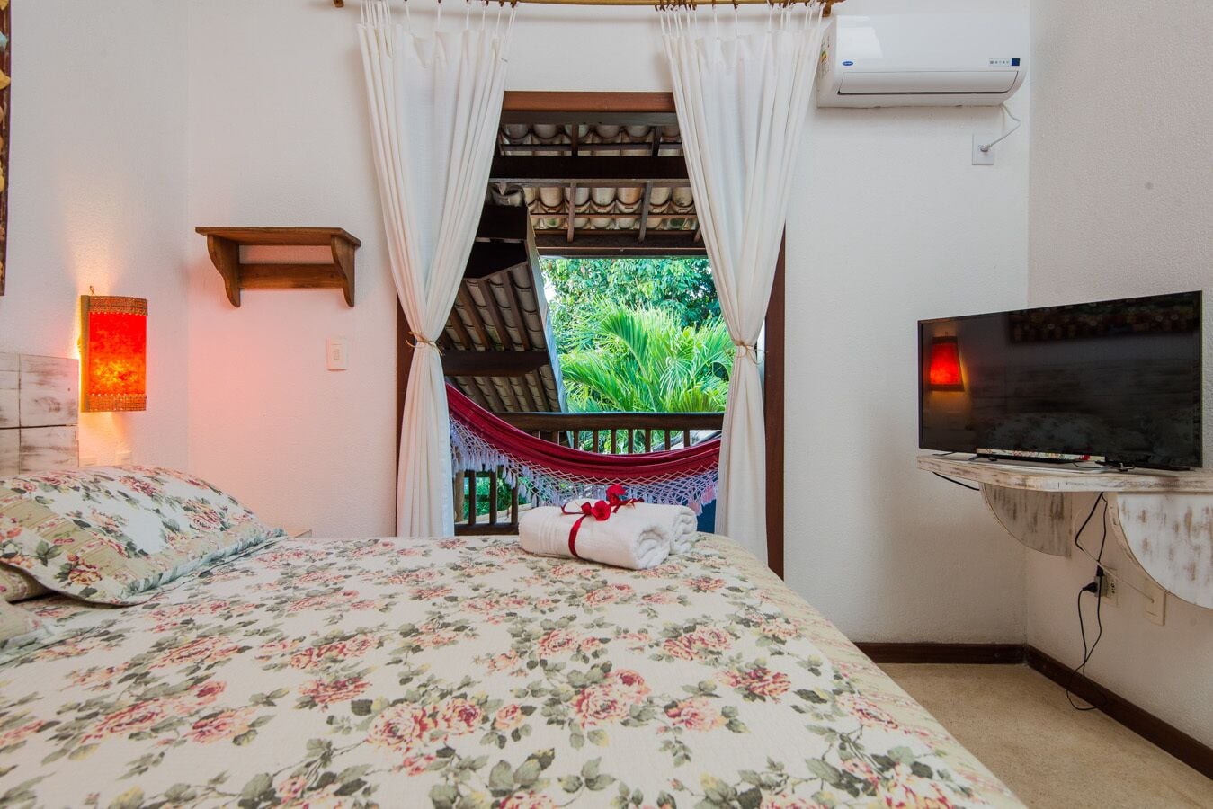 A bedroom with a bed, a TV, a hammock, and a view of greenery outside.