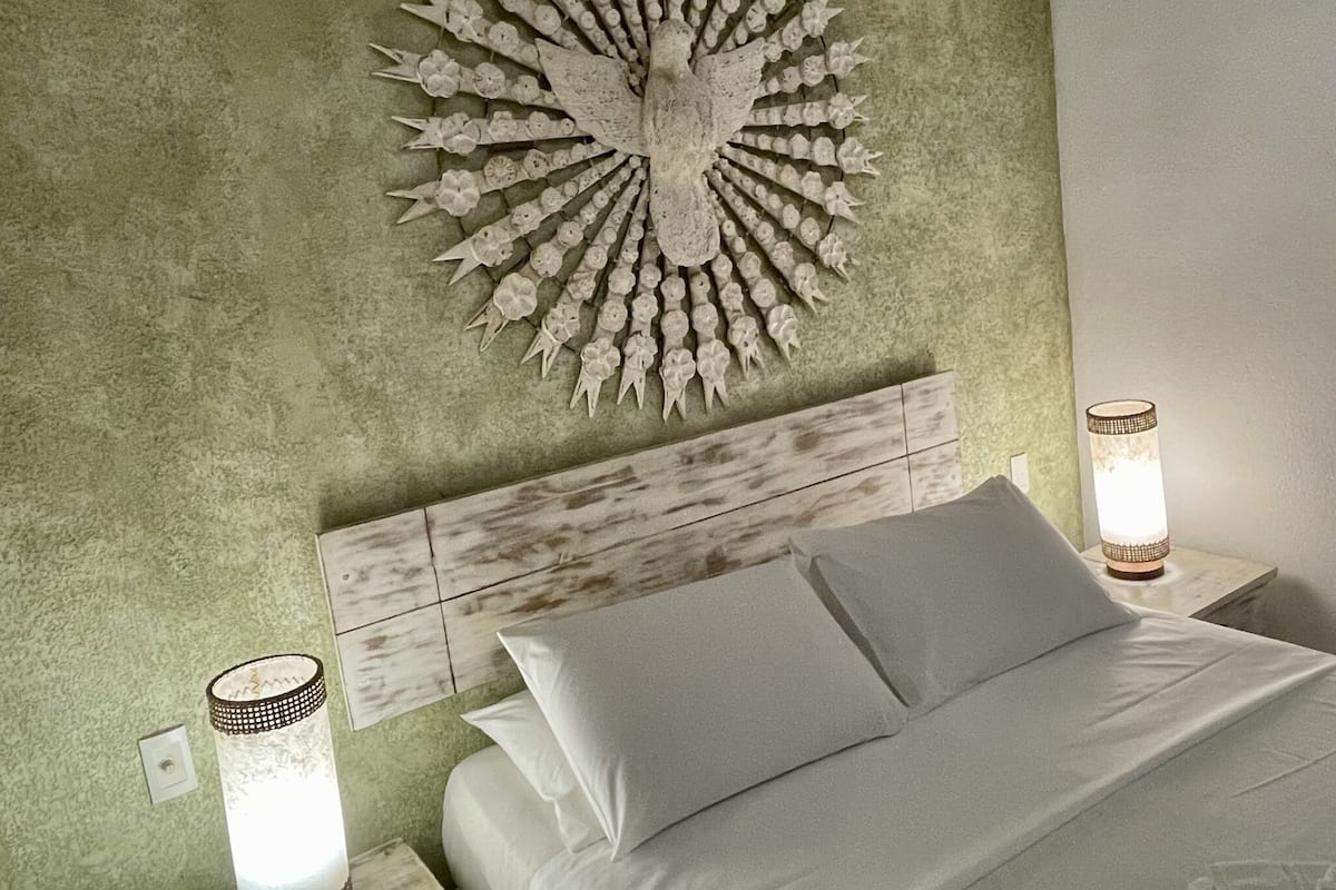 A bedroom with a green textured wall, a wooden headboard, a white bed with two pillows, and a wall-mounted light fixture.