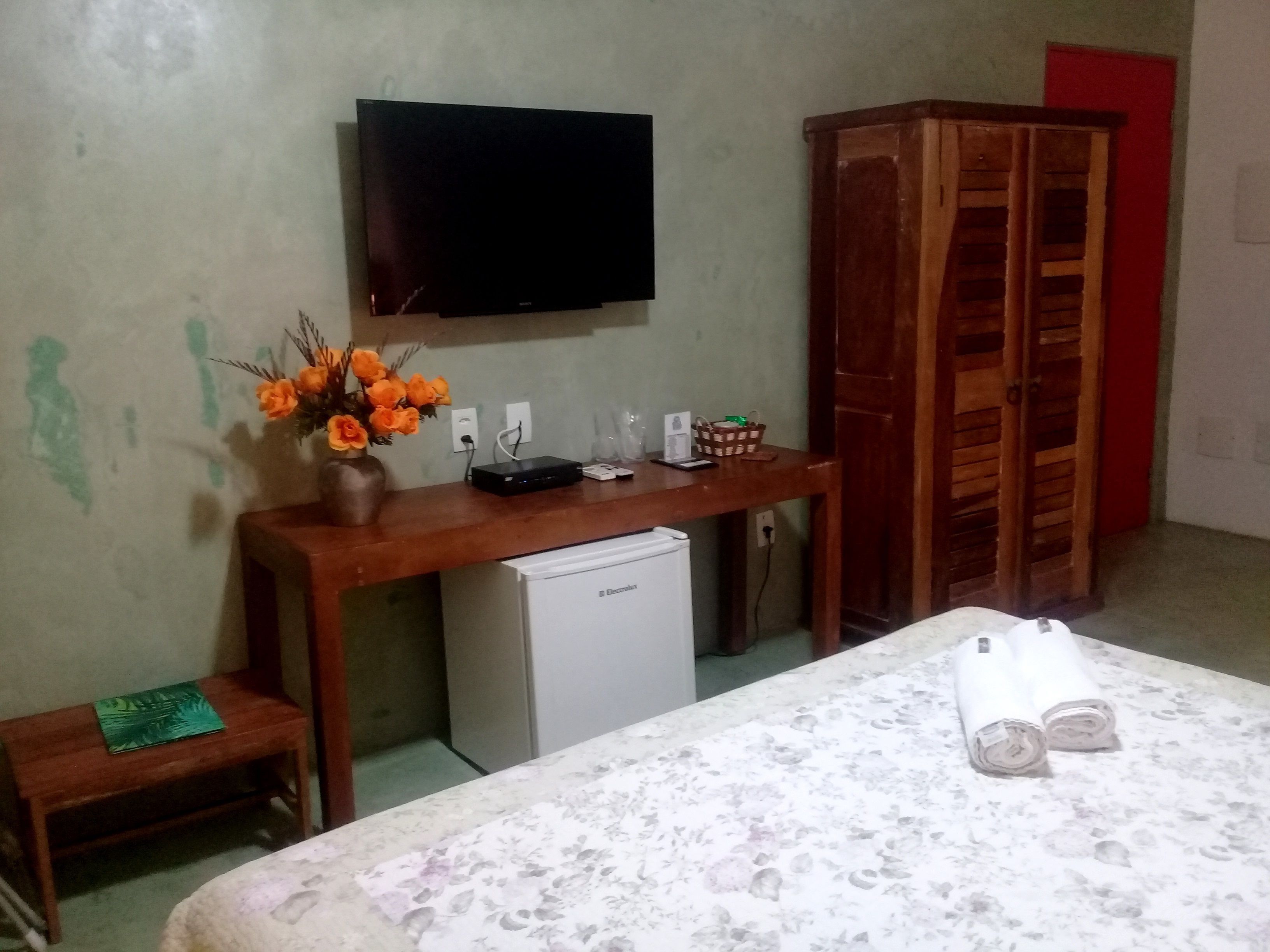 A room with a bed, a wooden desk with a television, a small refrigerator, and a wooden wardrobe.