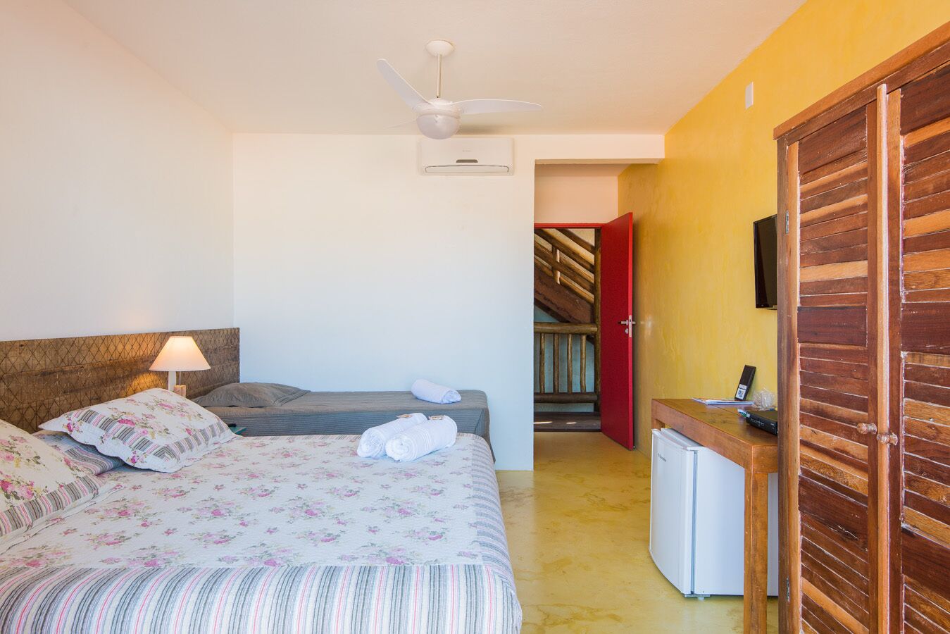 Un dormitorio con cama, escritorio, silla, lámpara, ventilador de techo y una puerta roja que da a una escalera.