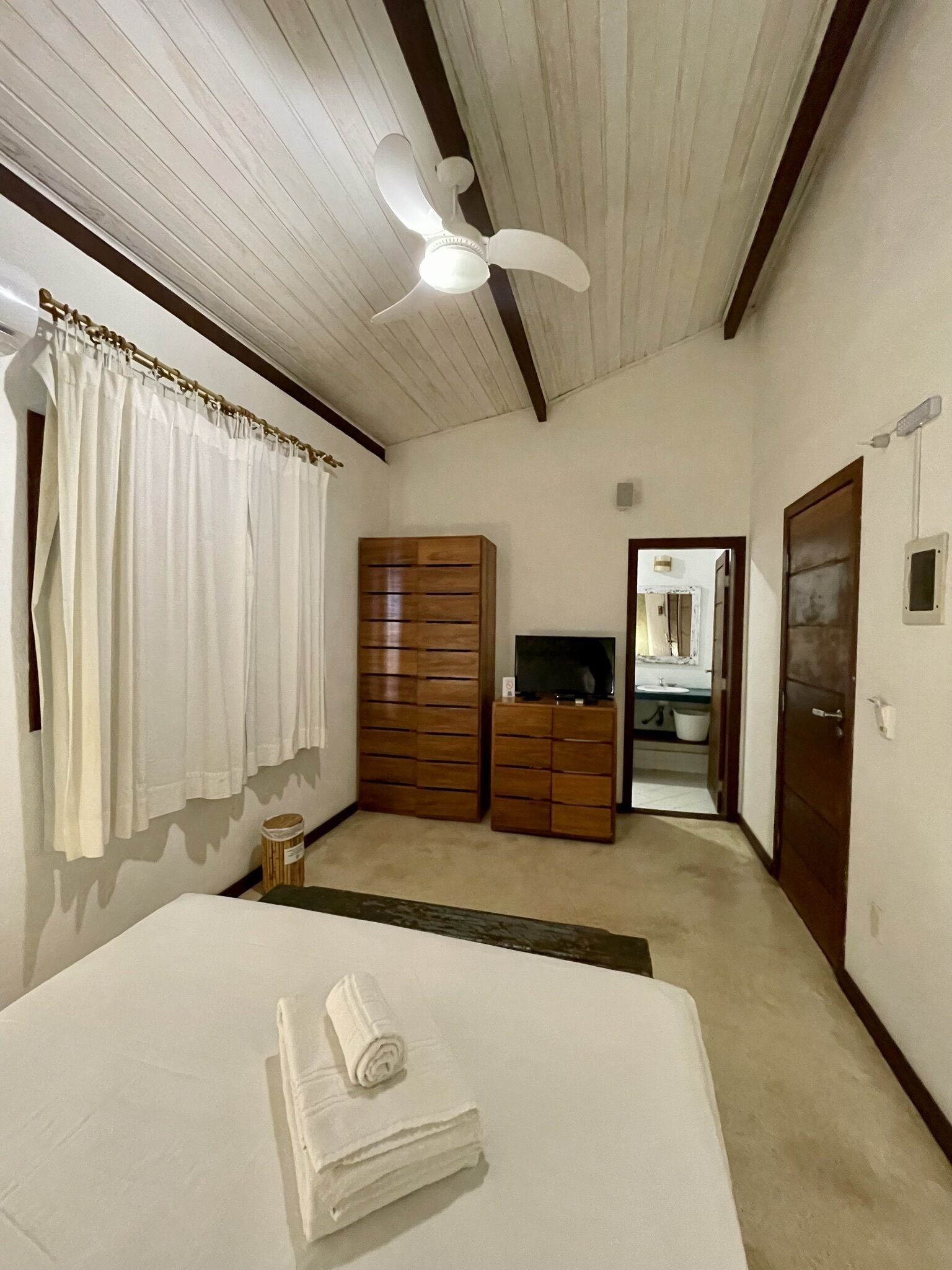 Un dormitorio con cama, ventilador de techo, armario de madera, televisión y baño visible a través de una puerta abierta.