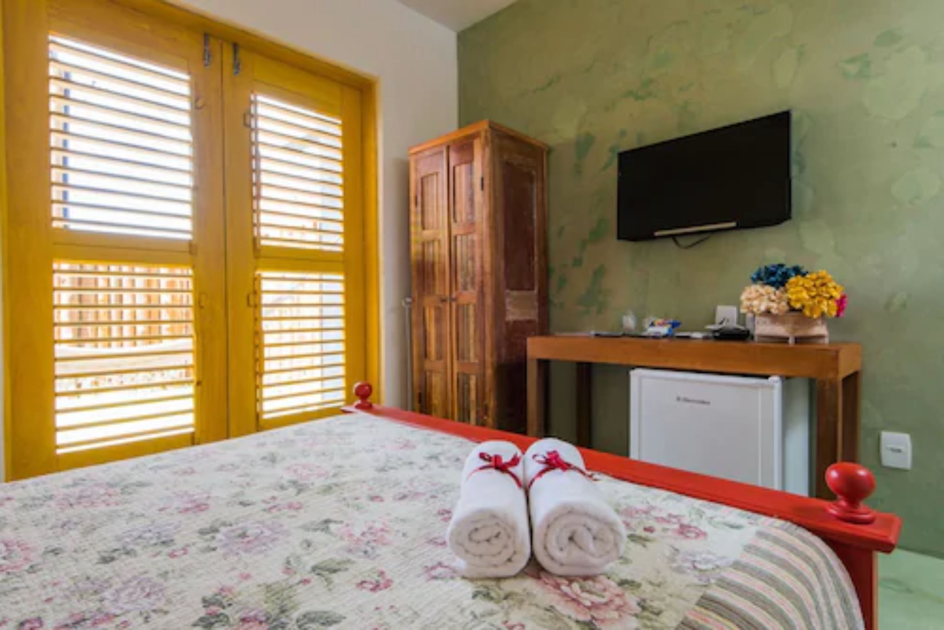 Quarto de hotel com cama, guarda-roupa de madeira, secretária pequena com televisão e frigorífico.