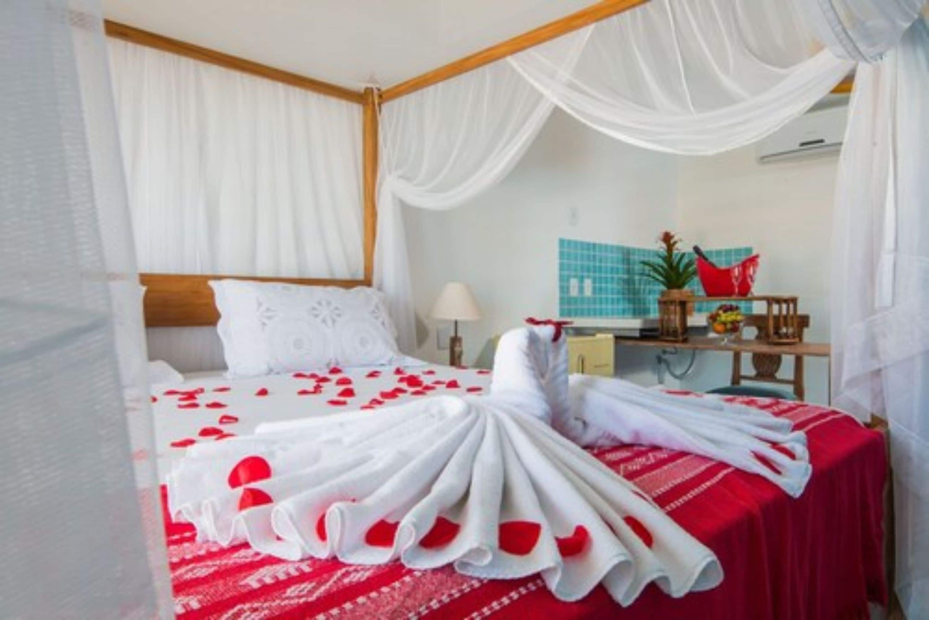 A hotel room with a bed covered in white linens and red rose petals, a wooden headboard, a bedside table with a lamp, and a small plant.
