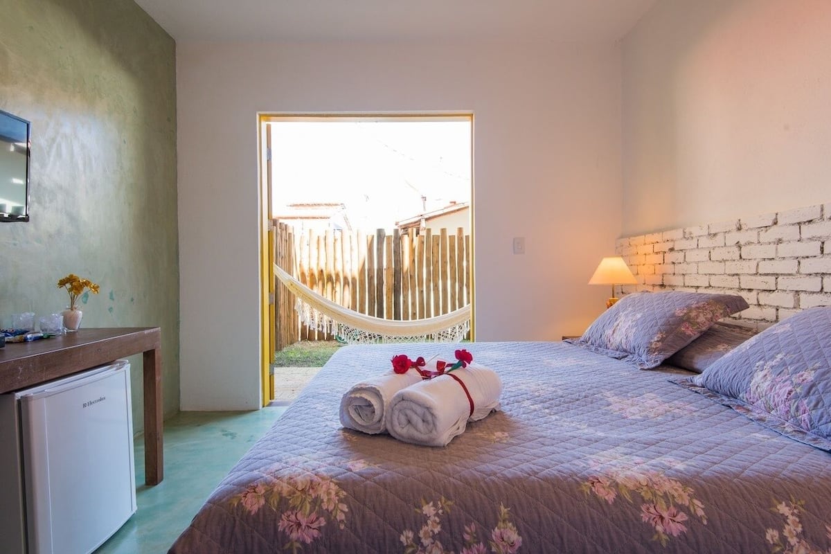 A bedroom with a bed, a desk, a hammock, and a view of the outside.