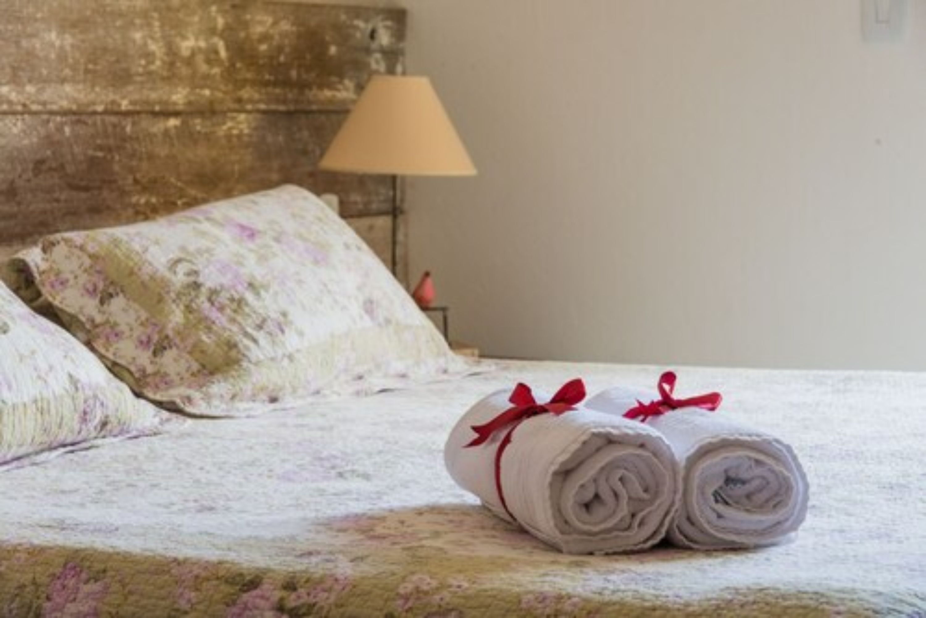 A neatly made bed with floral-patterned pillows and rolled white towels with red ribbons, set against a rustic wooden headboard and a lamp on a bedside table.