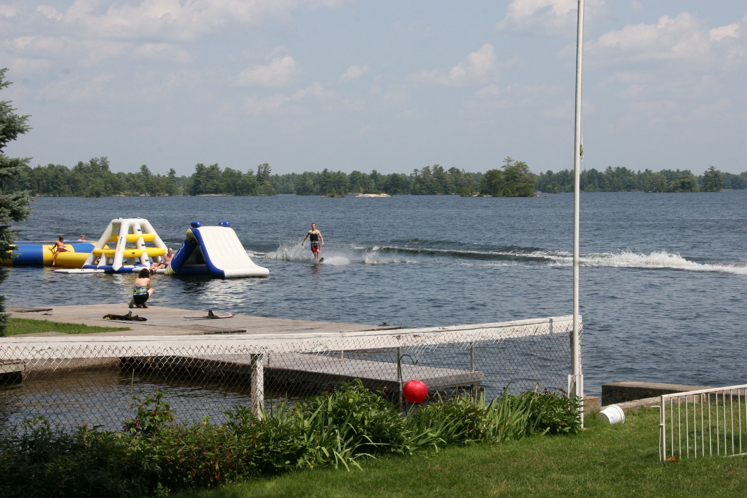 Une scène au bord d’un lac avec un toboggan aquatique, des structures de jeux gonflables et une personne pratiquant le ski nautique.