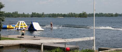 Une scène au bord d’un lac avec un toboggan aquatique, des structures de jeux gonflables et une personne pratiquant le ski nautique.
