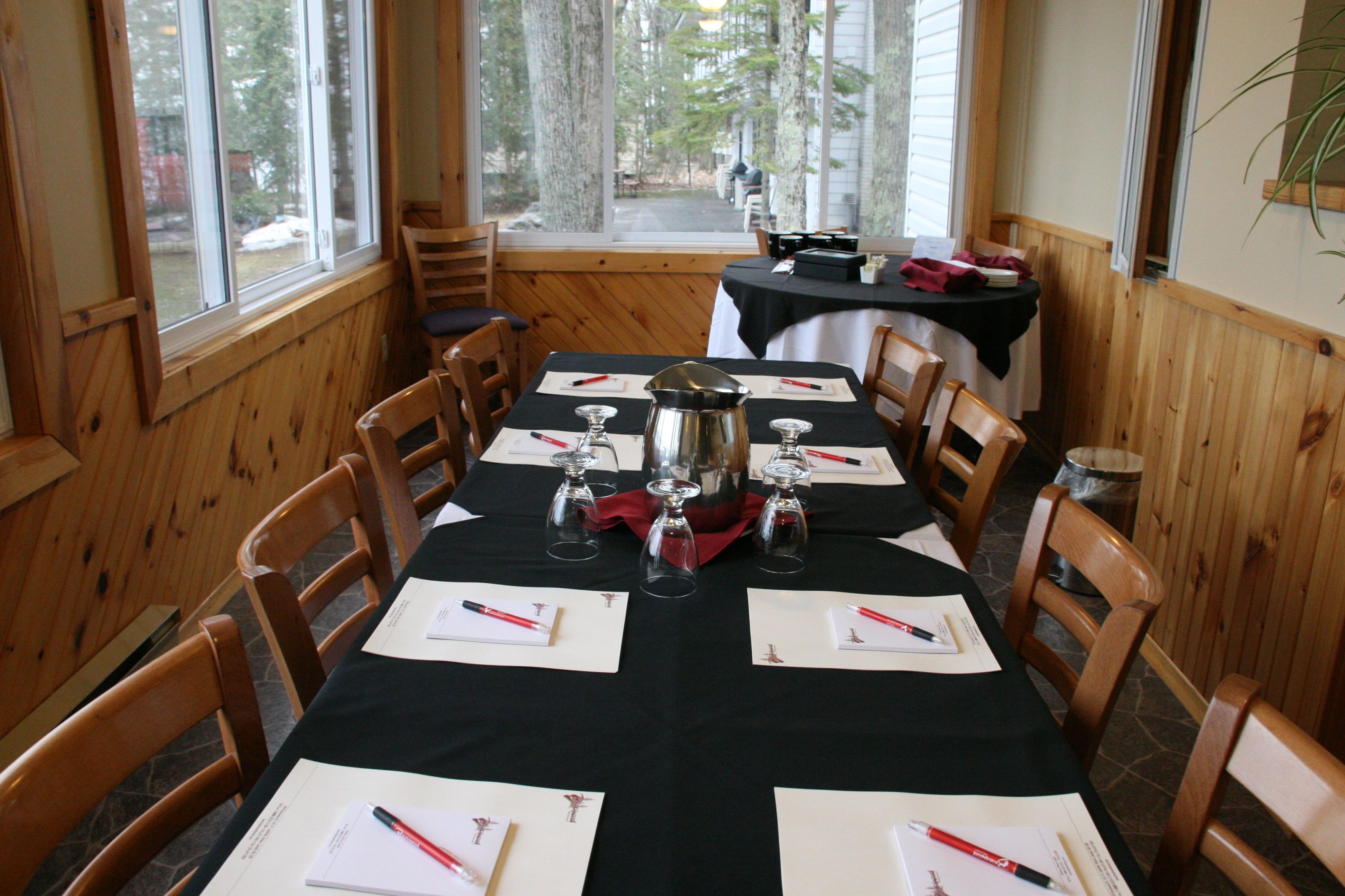 A dining area with a long table set for four, wooden chairs, and a view of trees through large windows.