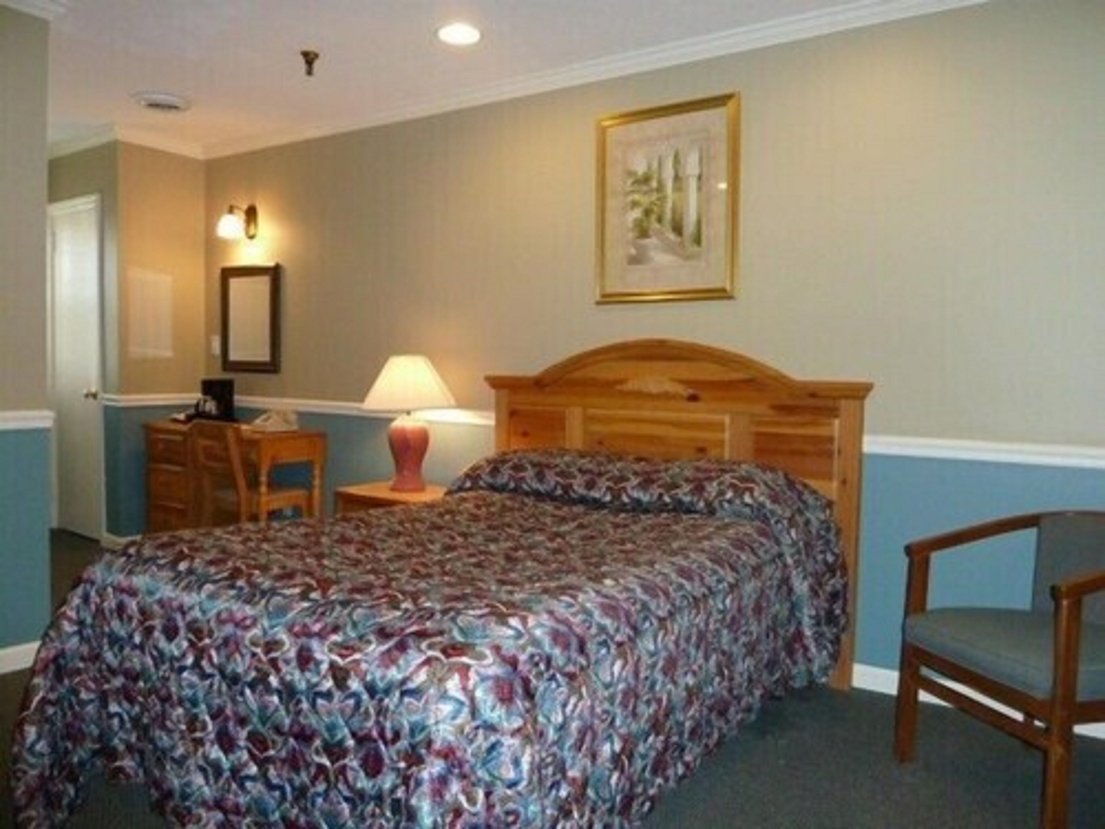A bedroom with a wooden headboard, a patterned bedspread, a chair, a desk with a lamp, and a framed picture on the wall.