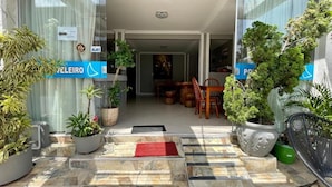 Entrada de hotel con un patio empedrado, plantas en macetas y un área de comedor visible en el interior.