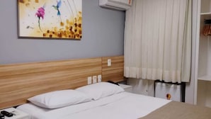 A hotel room with a wooden headboard, a bed with white linens, a nightstand with a phone and charger, a painting on the wall, and an air conditioning unit. - Hotel Três Caravelas (Fortaleza)