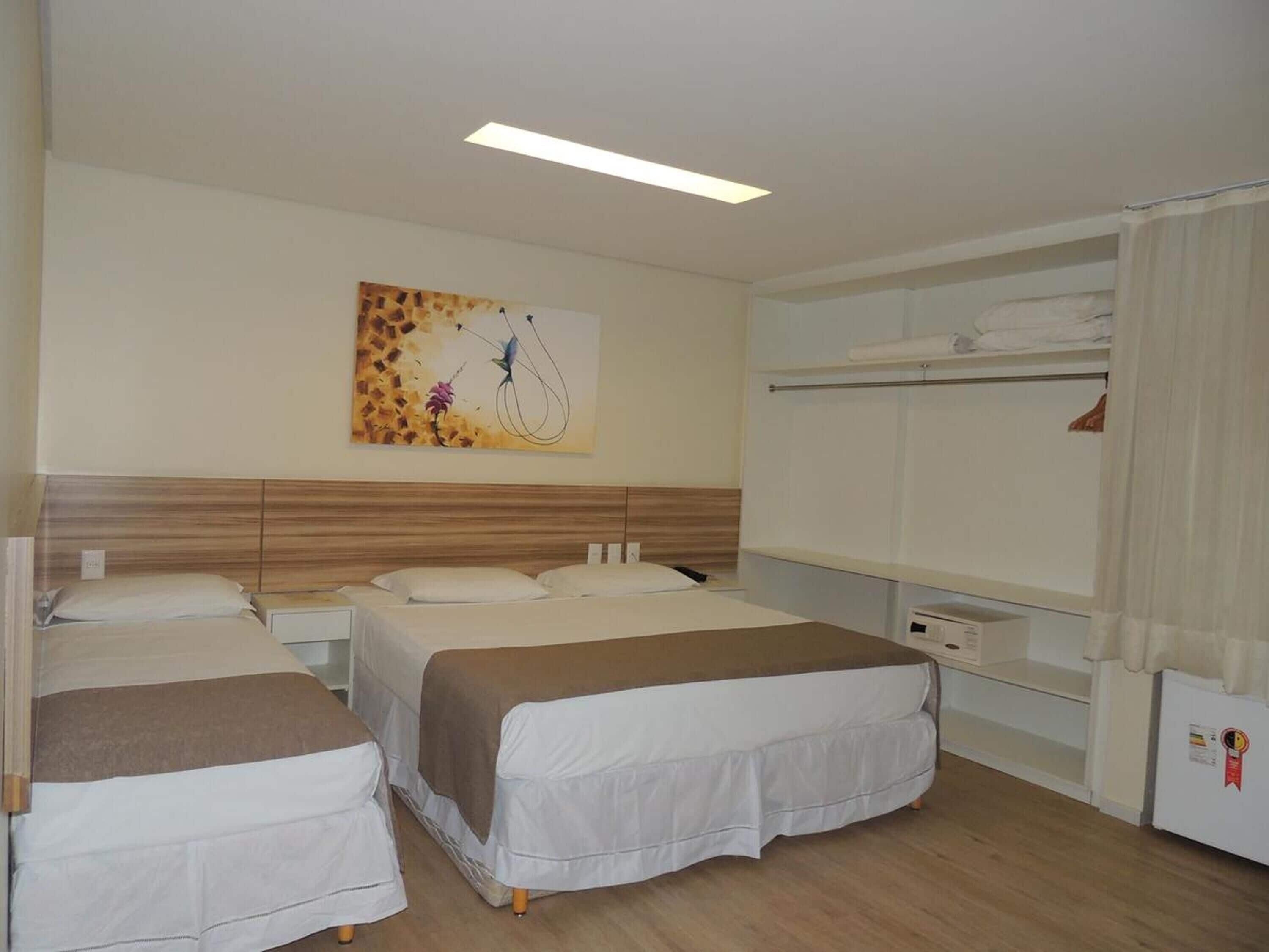A hotel room with two beds, a wooden headboard, a painting on the wall, a wardrobe, and a small refrigerator.