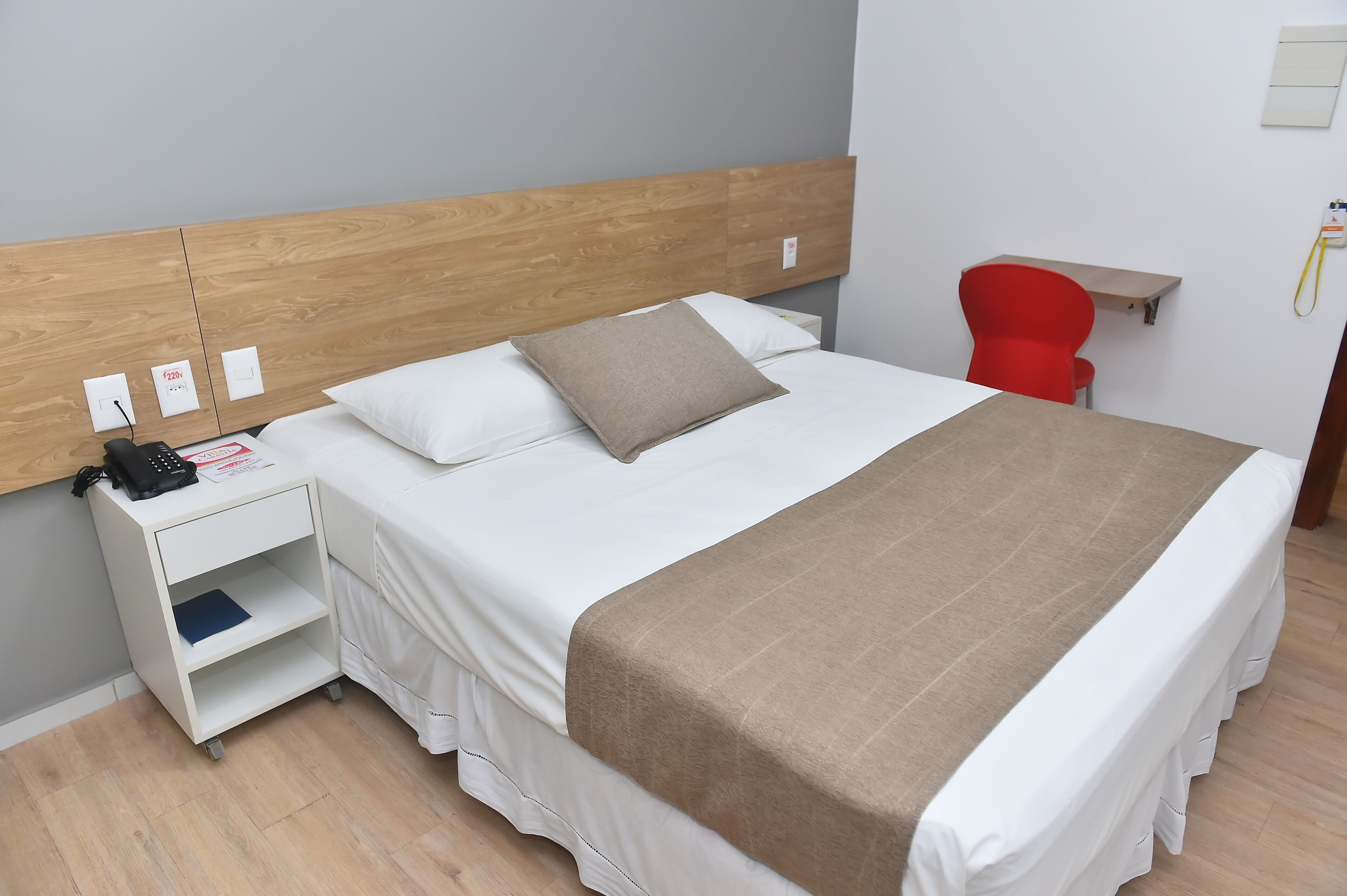 Una cama bien hecha con cabecera de madera, una mesita de noche con un teléfono y un libro, y una silla roja en una habitación sencilla con paredes y suelo de colores claros.