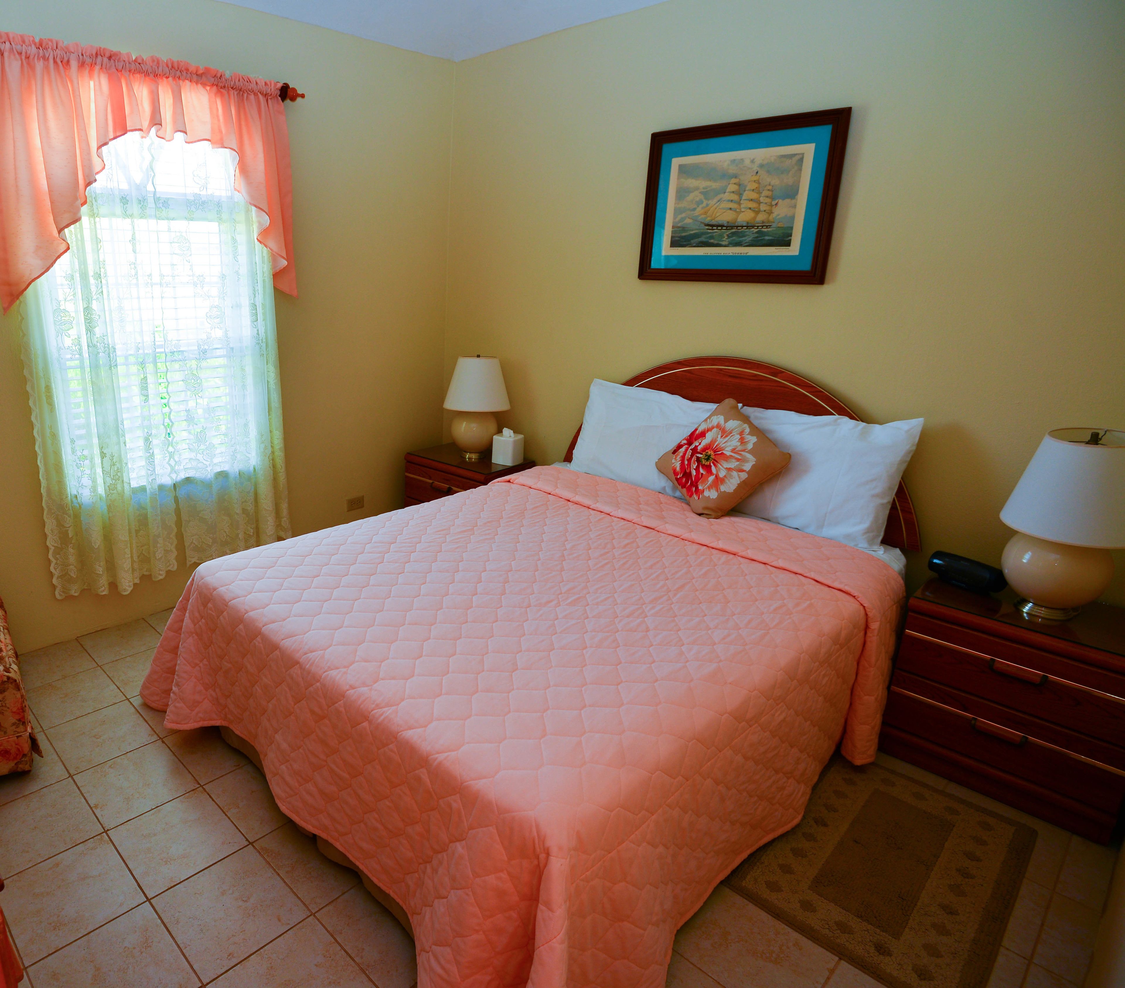 A neatly made bed with a pink quilt, a bedside lamp, a nightstand, a framed picture on the wall, and a window with curtains.