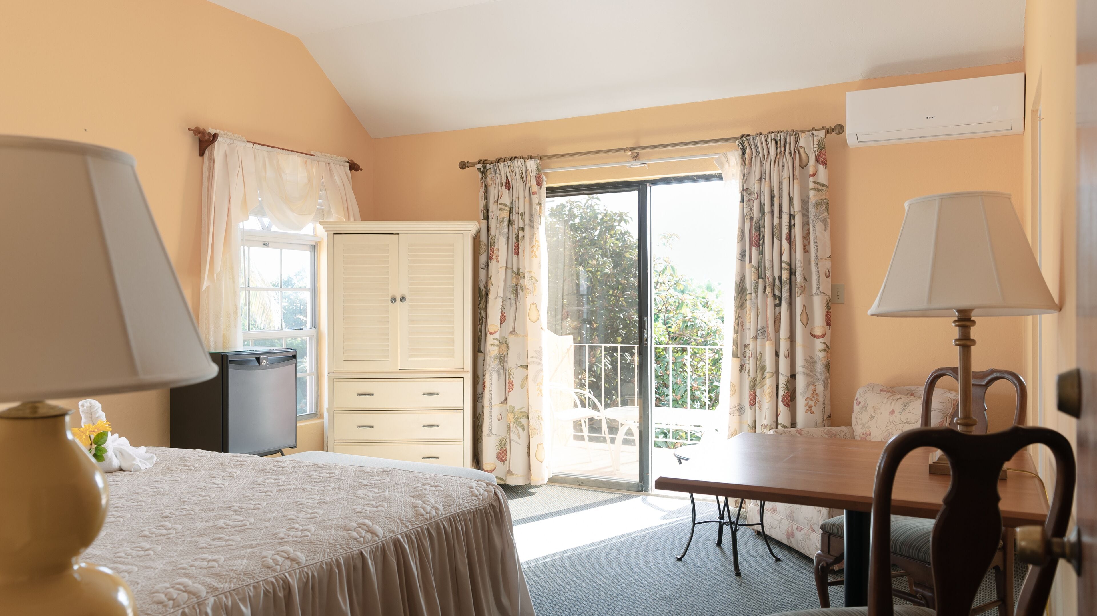 A hotel room with a bed, a desk, a chair, a window with curtains, a balcony, and an air conditioning unit.
