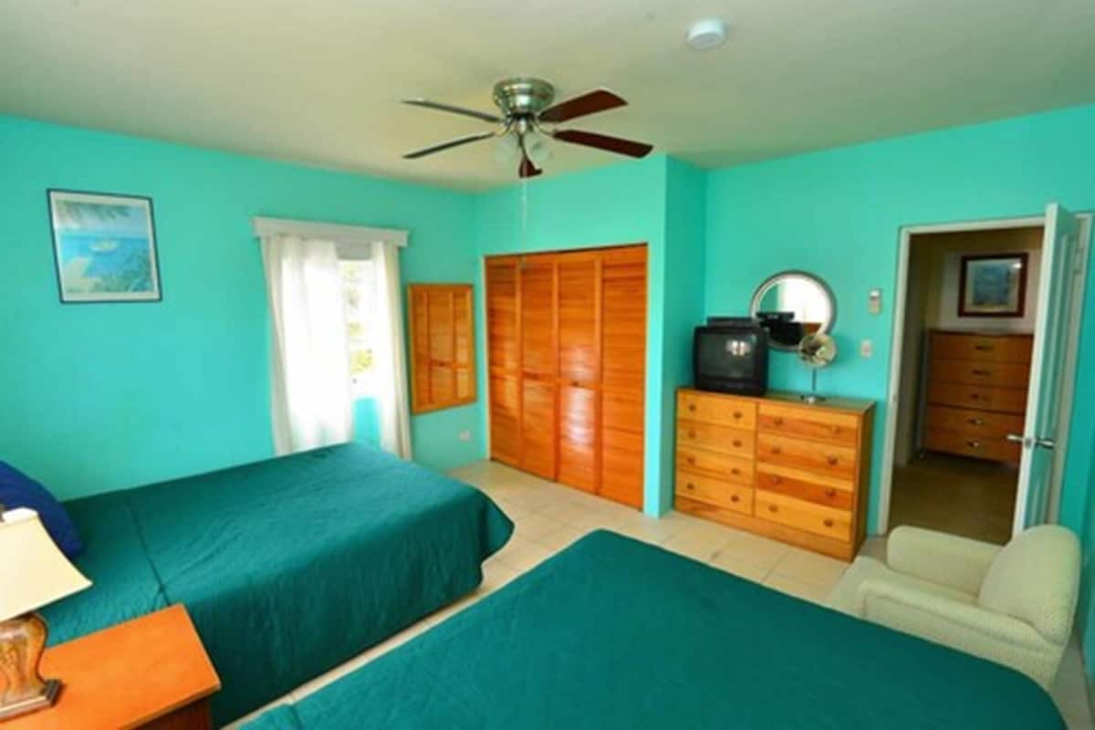 Quarto com duas camas, ventilador de teto, guarda-roupa de madeira, cômoda e televisão.