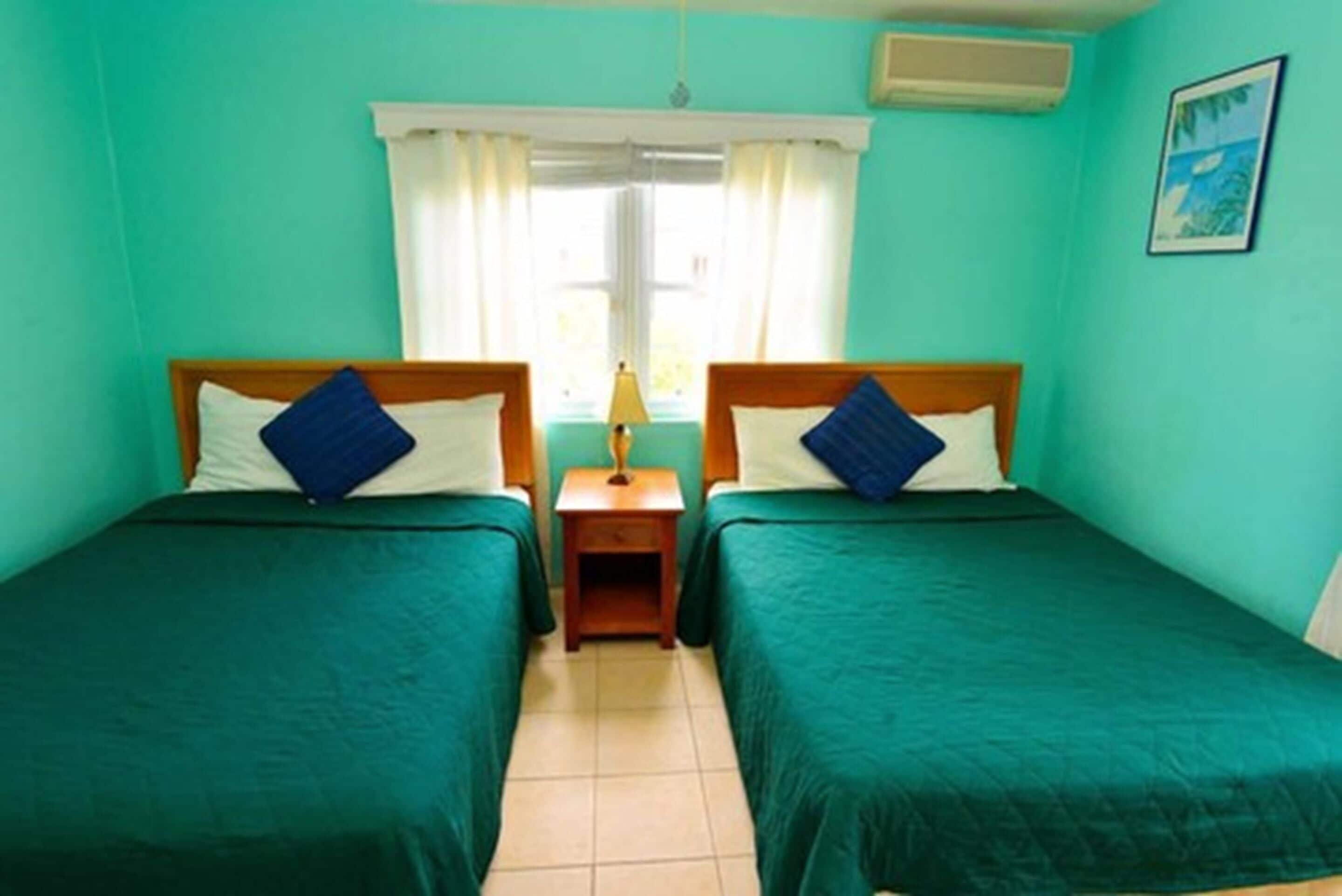 Une chambre avec deux lits, chacun doté d’une tête de lit en bois et d’une literie verte. Une petite table de chevet avec une lampe se trouve entre les deux lits.