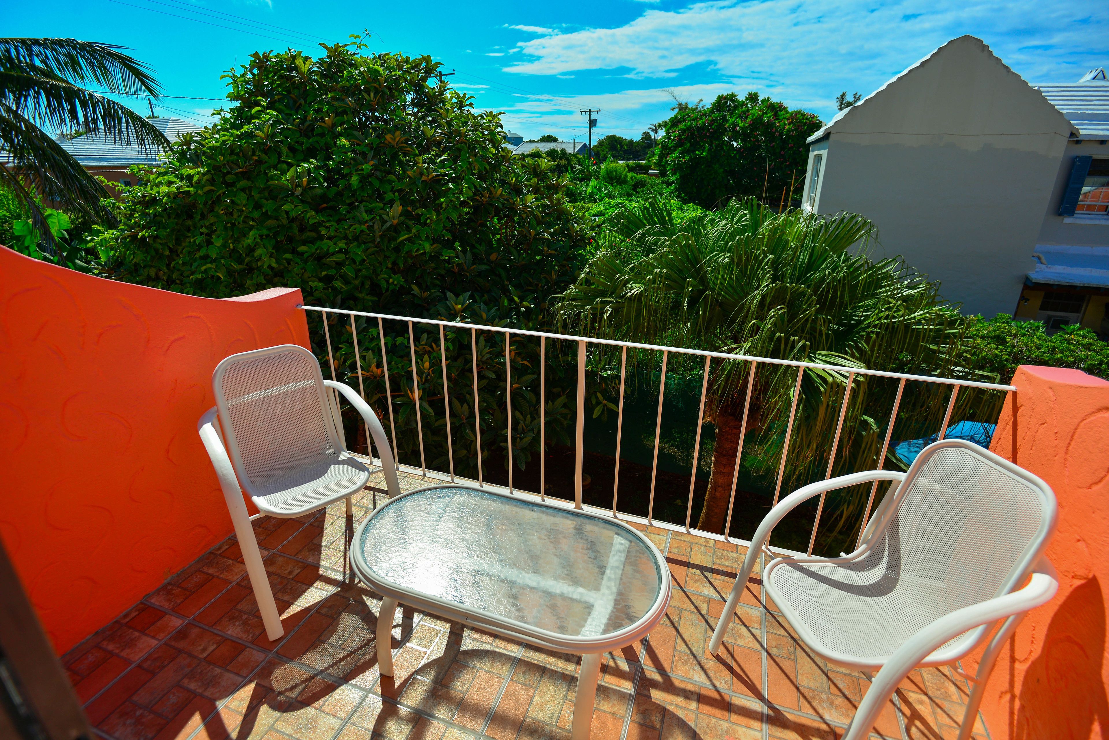 A balcony with two white chairs and a glass table, surrounded by orange walls and a white railing.