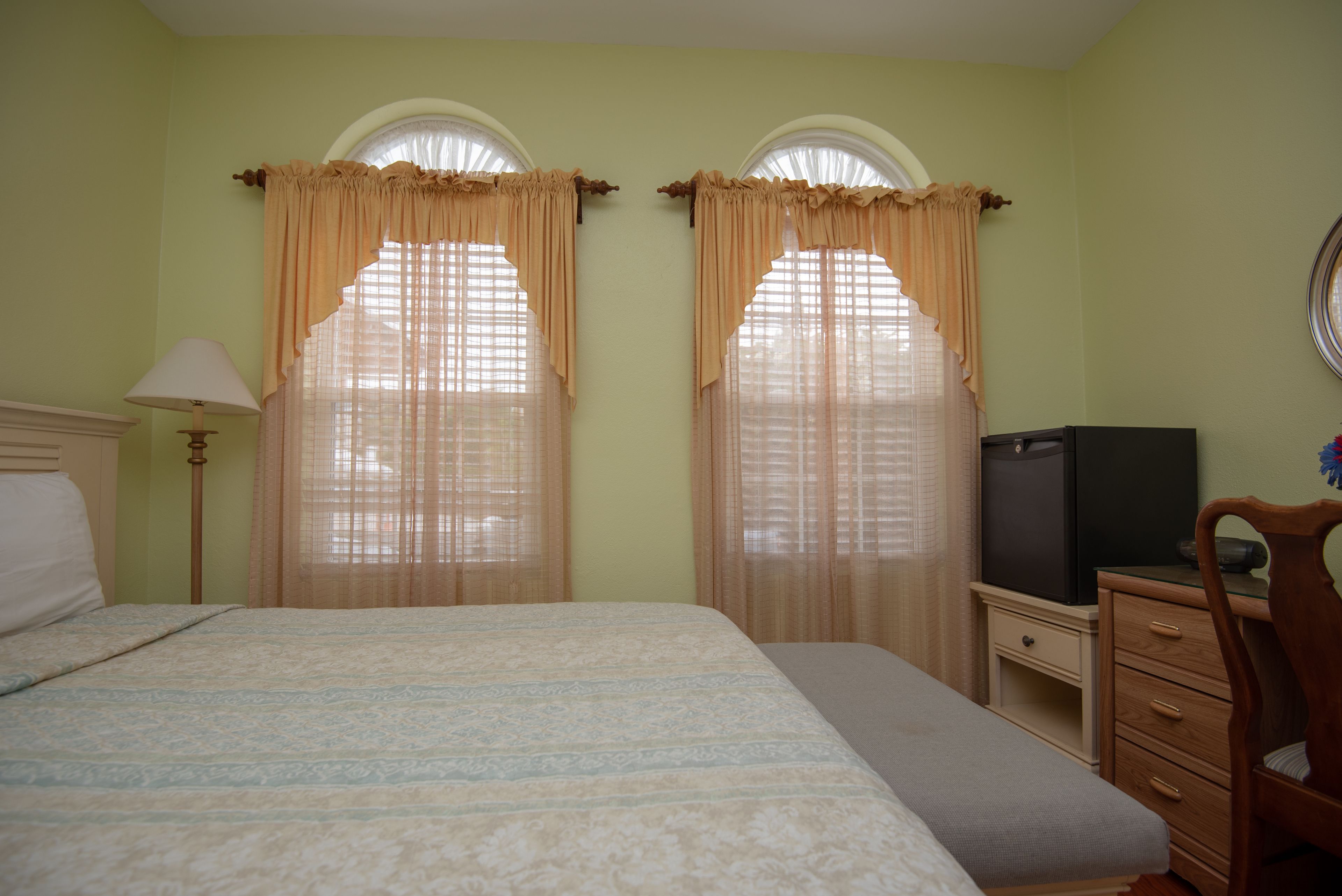 A hotel room with a bed, a desk with a television, and two windows with curtains.