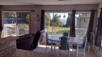 A spacious room with a large sliding glass door leading to a balcony. Inside, there is a dining table with chairs, a black leather sofa, and a marble countertop.