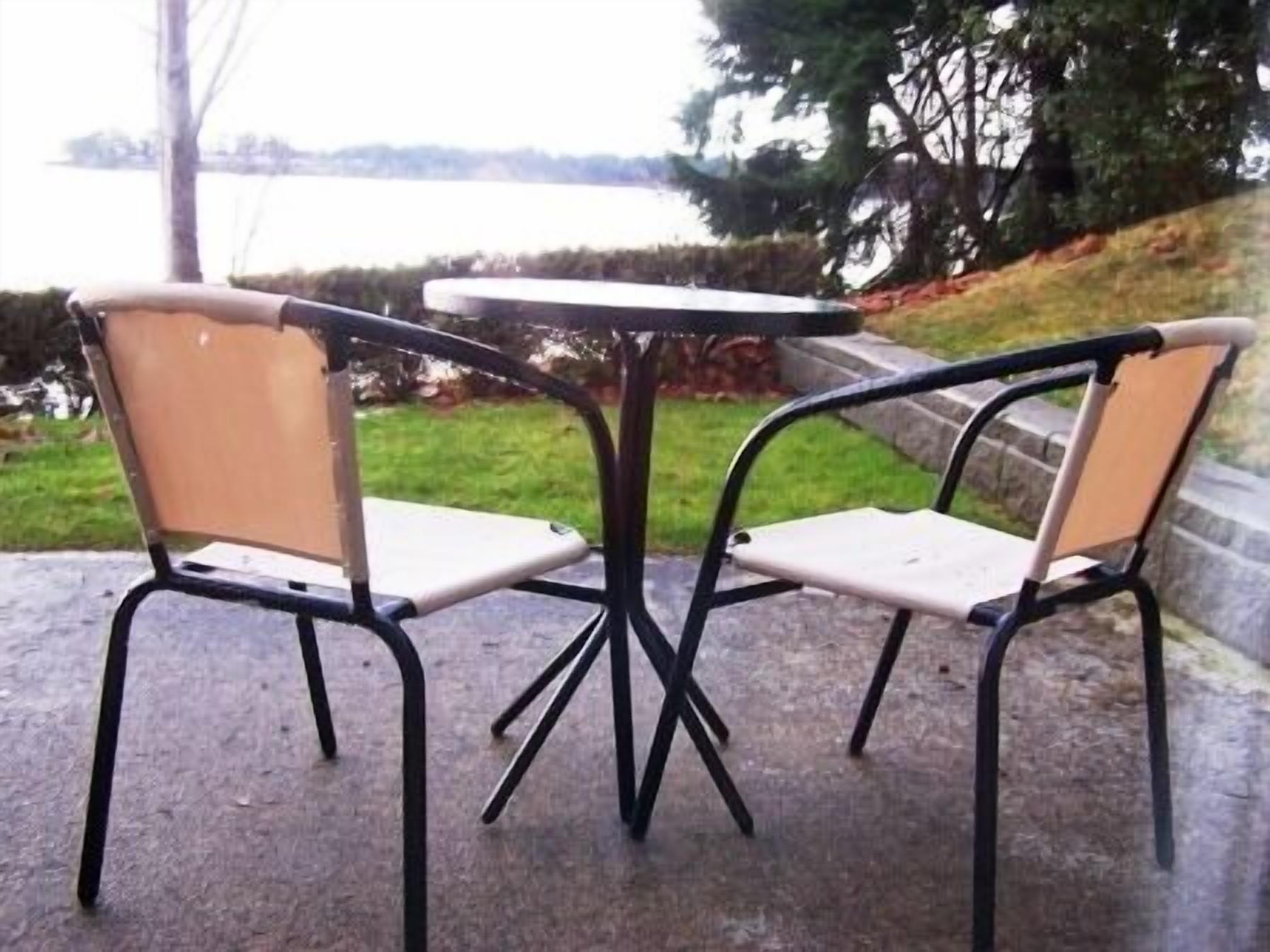 Dos sillas con una mesa en medio, sobre una superficie de concreto, con vista a un cuerpo de agua y árboles al fondo.