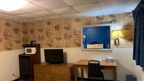 A hotel room with a wooden desk, a television, a small cabinet, a chair, a trash can, and a window with a view of a blue sky and a building.