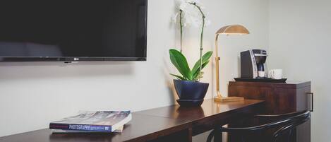 Una habitación moderna con una televisión de pantalla plana montada en la pared, un escritorio de madera con un libro, una orquídea en maceta, una lámpara y una cafetera.
