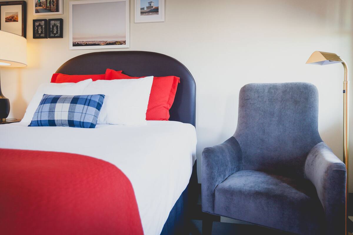 Hotellihuone, jossa on sänky, harmaa nojatuoli, lamppu ja seinällä olevia kehystettyjä kuvia.