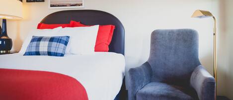 Una habitación de hotel con una cama, un sillón gris, una lámpara y cuadros en la pared.