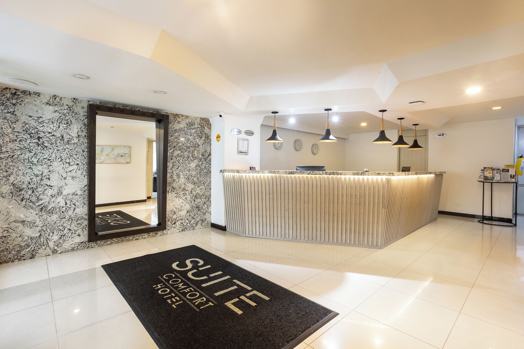 A hotel reception area with a marble wall, a reception desk, and a black doormat with 'SUITE COMFORT HOTEL' written on it.