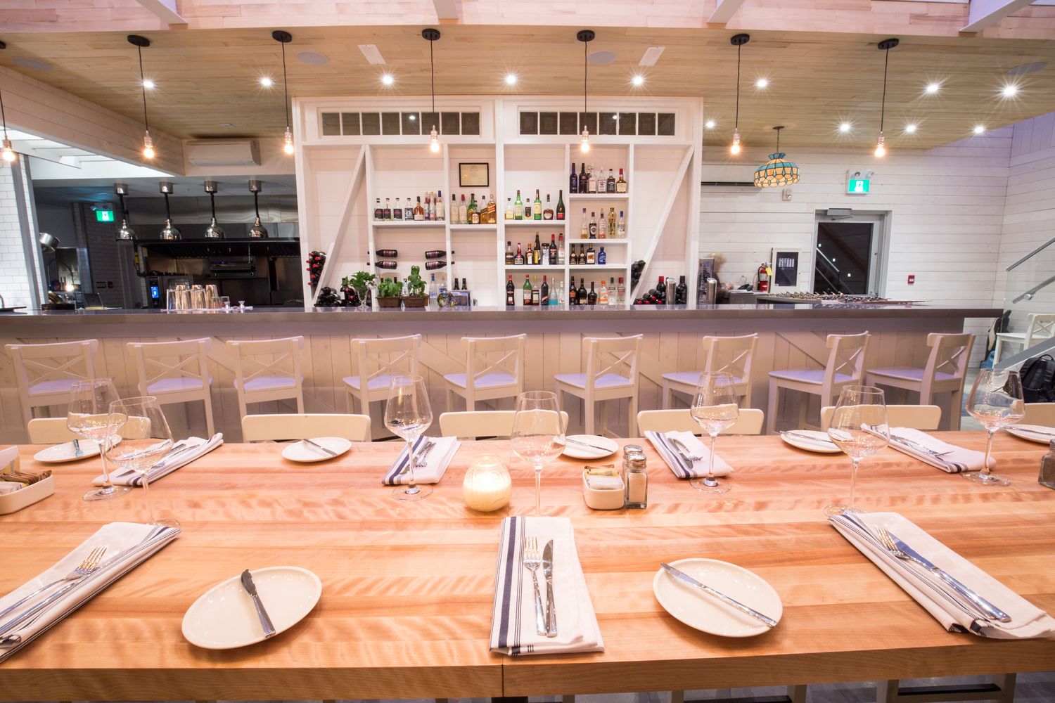 Un restaurante con mesas y sillas de madera, una zona de bar con estantes llenos de botellas y lámparas colgantes.