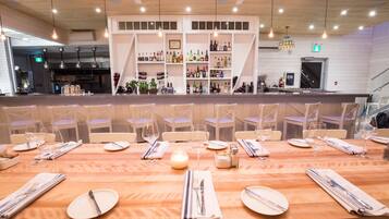 Un restaurante con mesas y sillas de madera, una zona de bar con estantes llenos de botellas y lámparas colgantes.