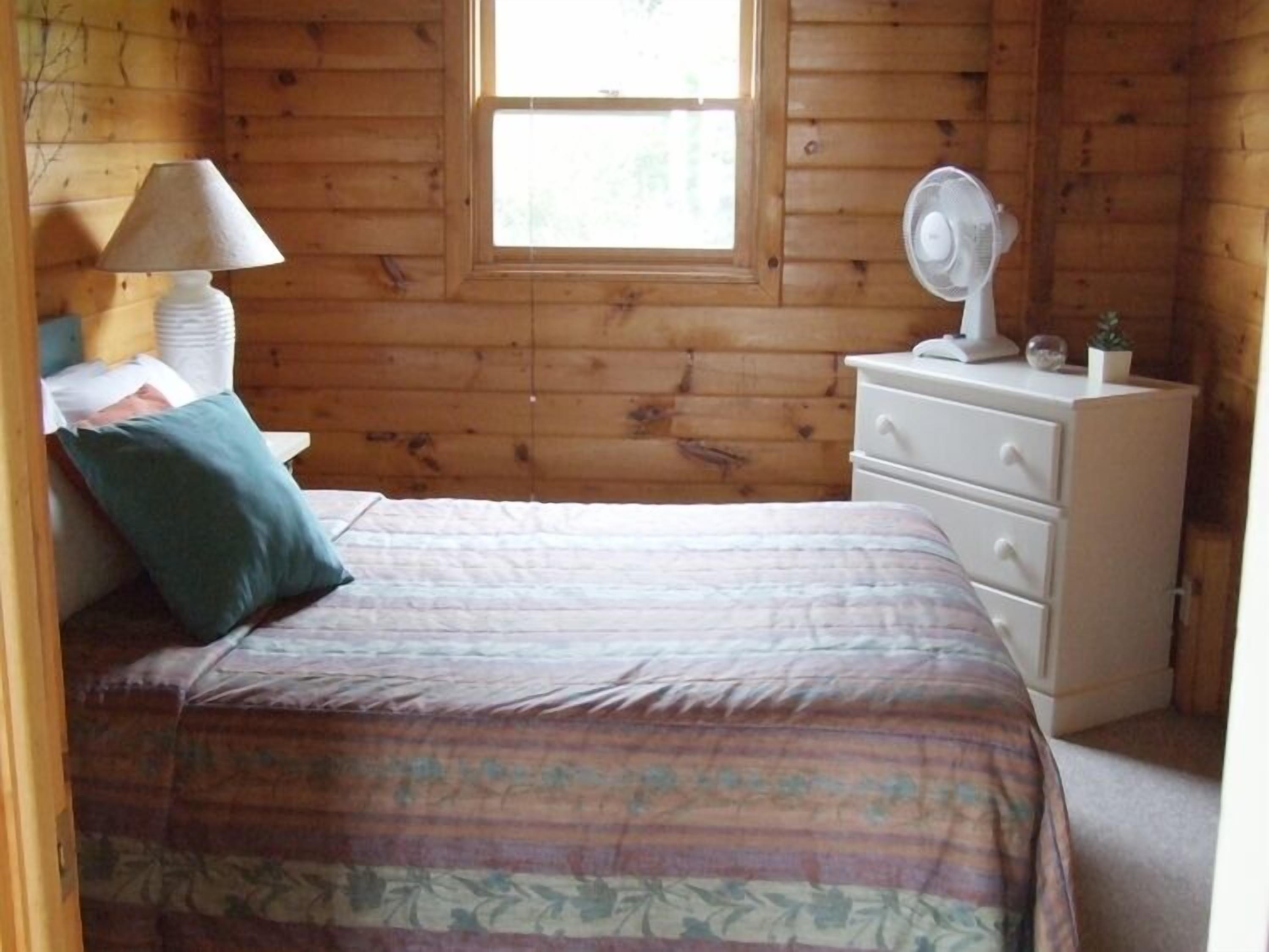 A wooden cabin room with a bed, a nightstand, a dresser, a lamp, and a fan.