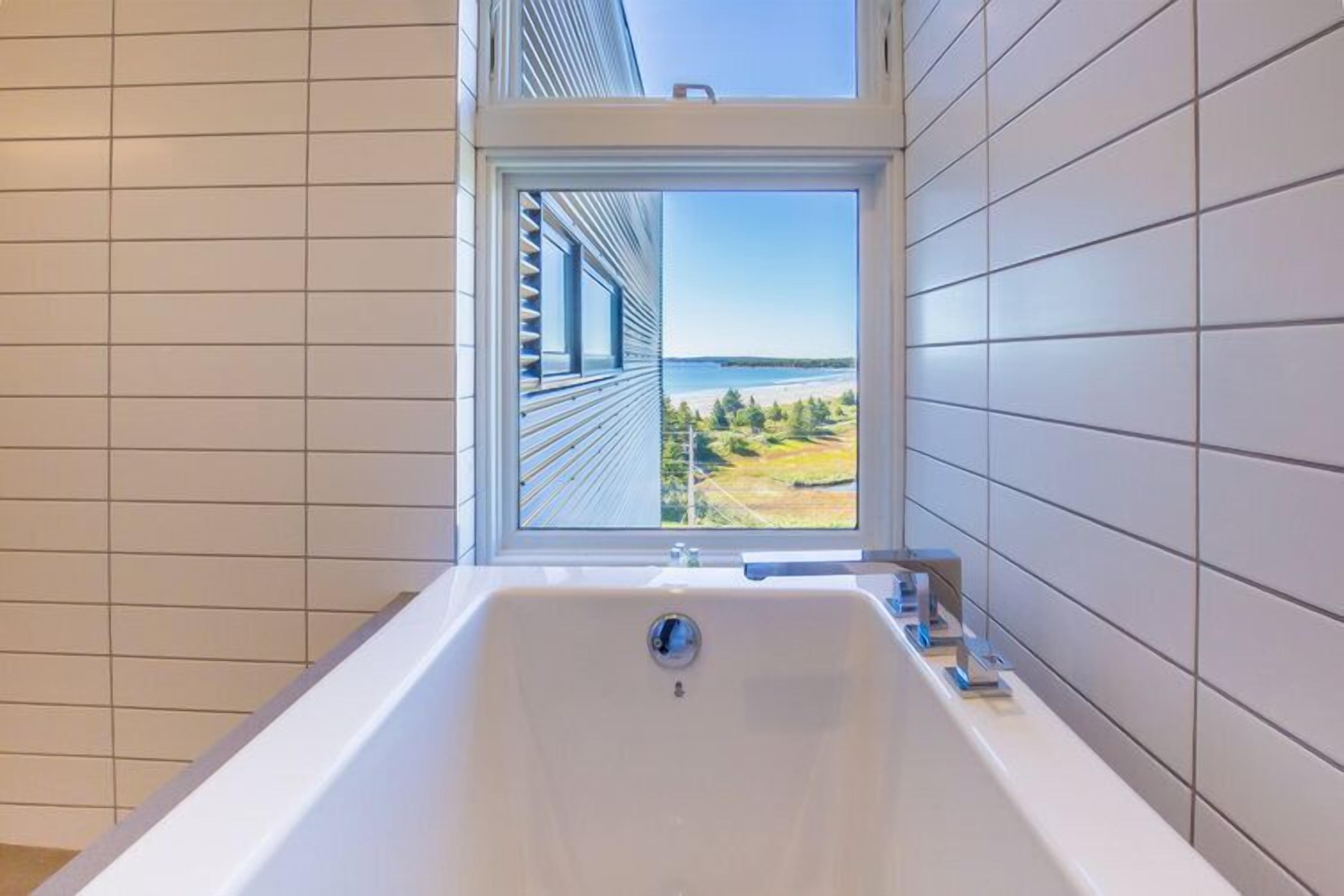 A bathroom with a large bathtub, a window with a view of a body of water and greenery, and tiled walls.