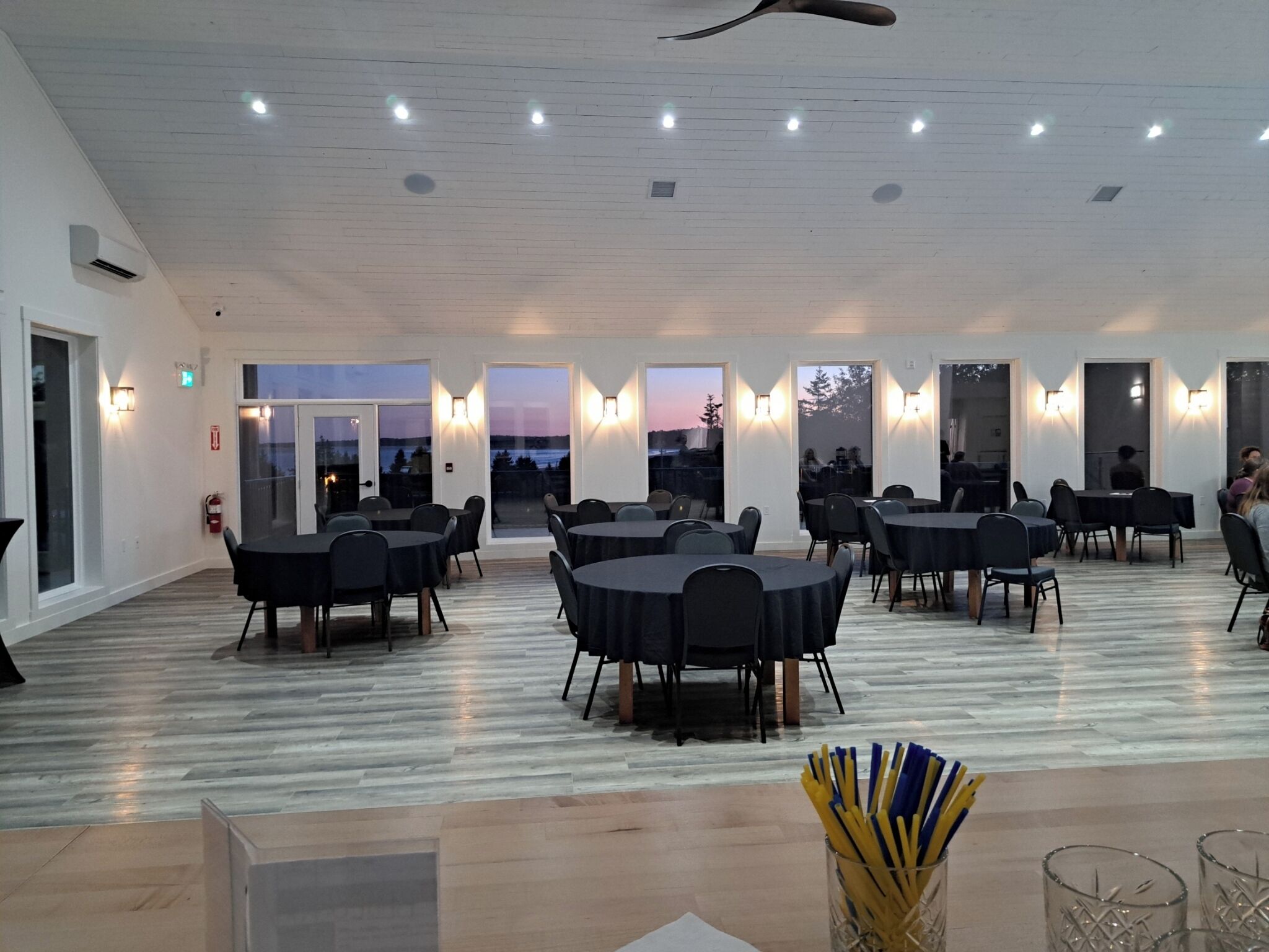 A spacious indoor area with multiple round tables and chairs, large windows with views, and a ceiling fan.