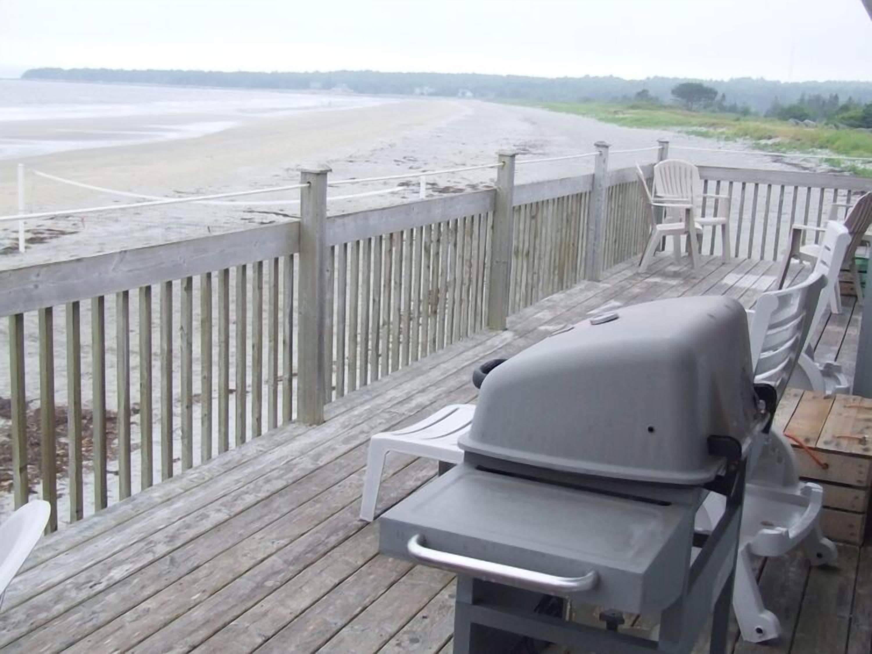 A wooden deck with a barbecue grill and white plastic chairs, overlooking a beach.