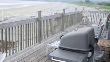 A wooden deck with a barbecue grill and white plastic chairs, overlooking a beach.