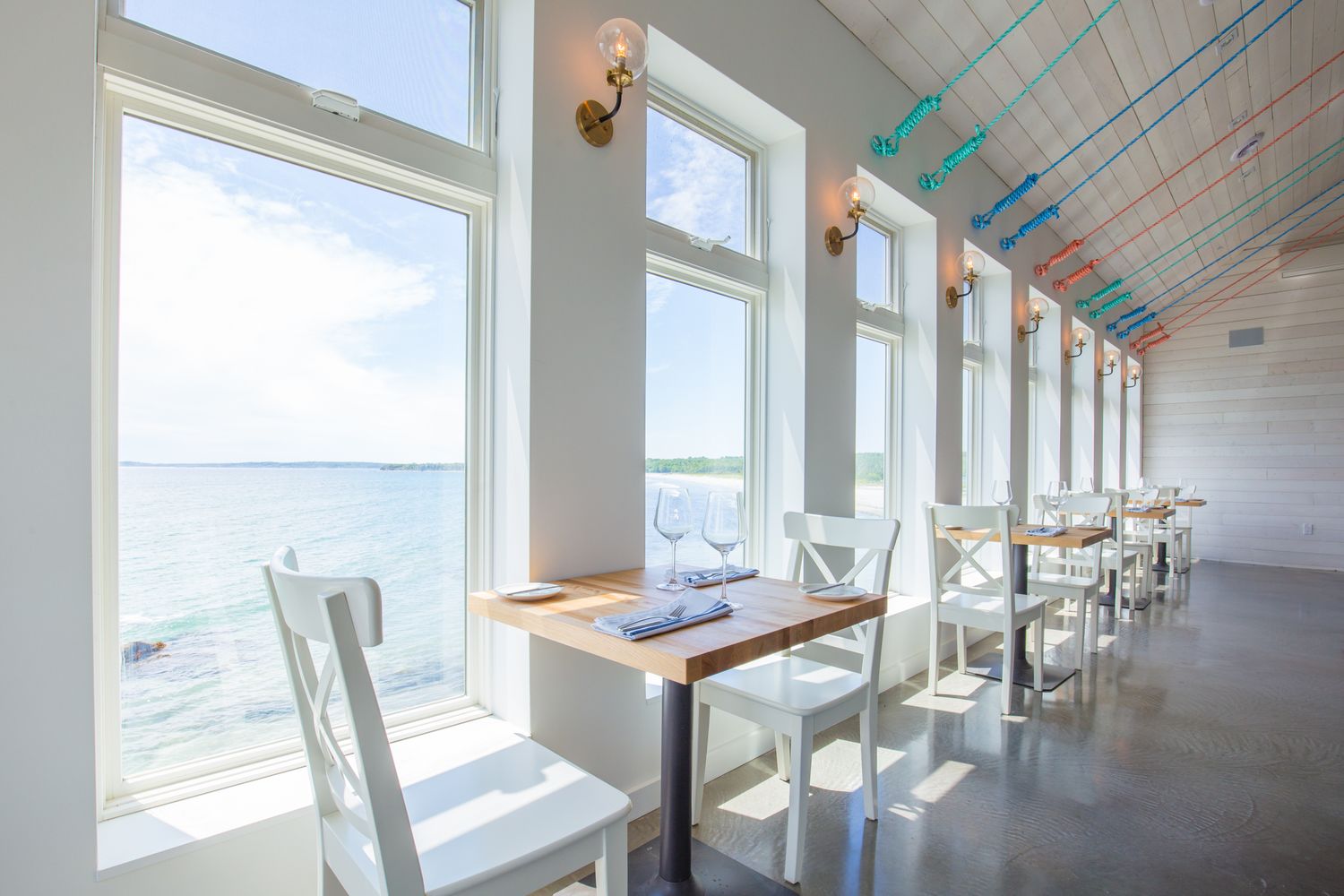 A restaurant with large windows offering a view of the sea. Tables are arranged with white chairs.