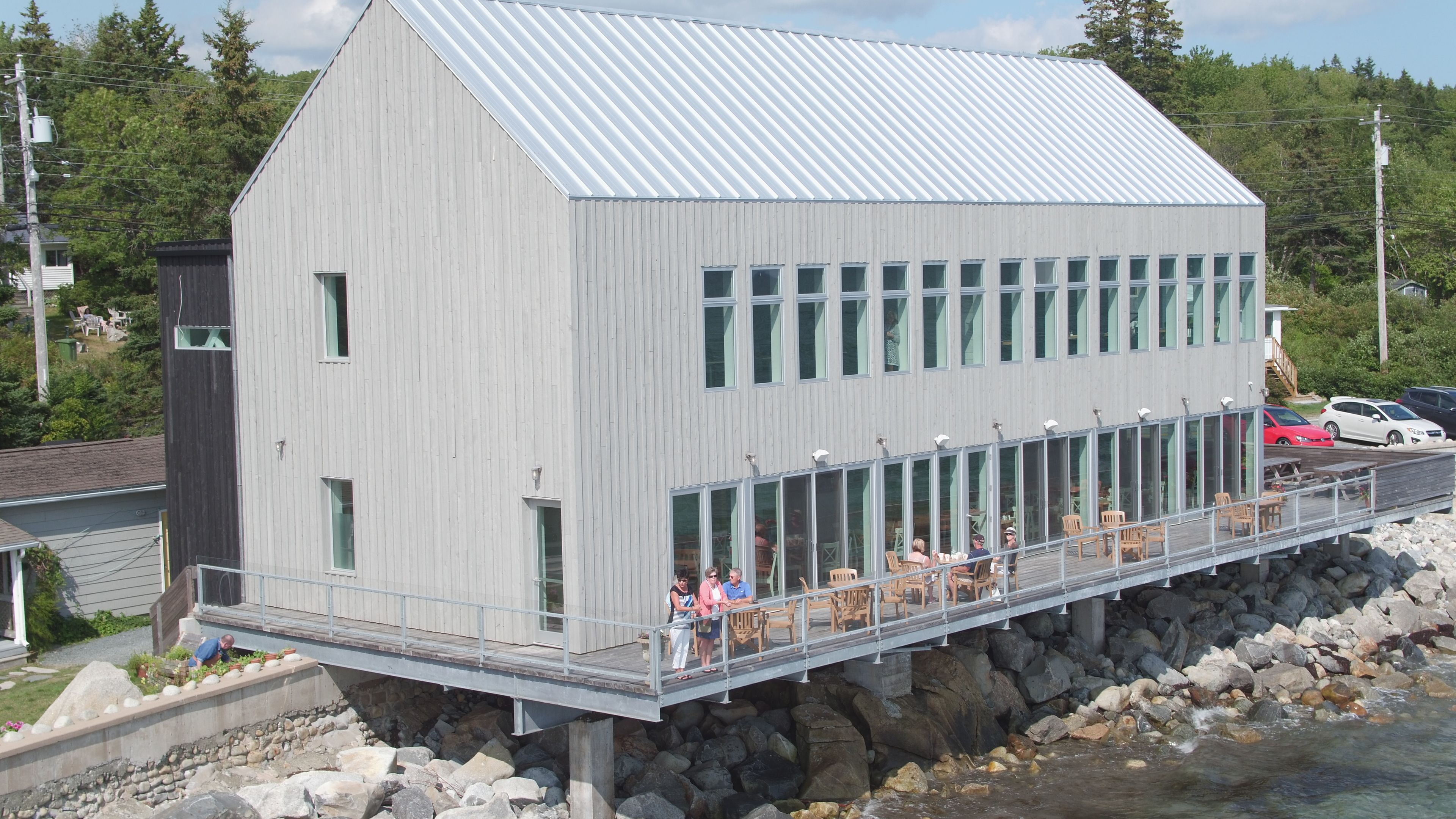A two-story building with a metal roof, a deck with outdoor seating, and a rocky shoreline.