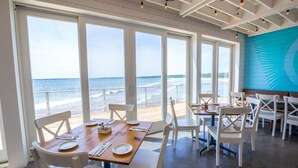 A dining area with large windows overlooking the ocean, wooden tables, white chairs, and a blue wall with a geometric design. - The Quarterdeck Resort (Summerville Centre)