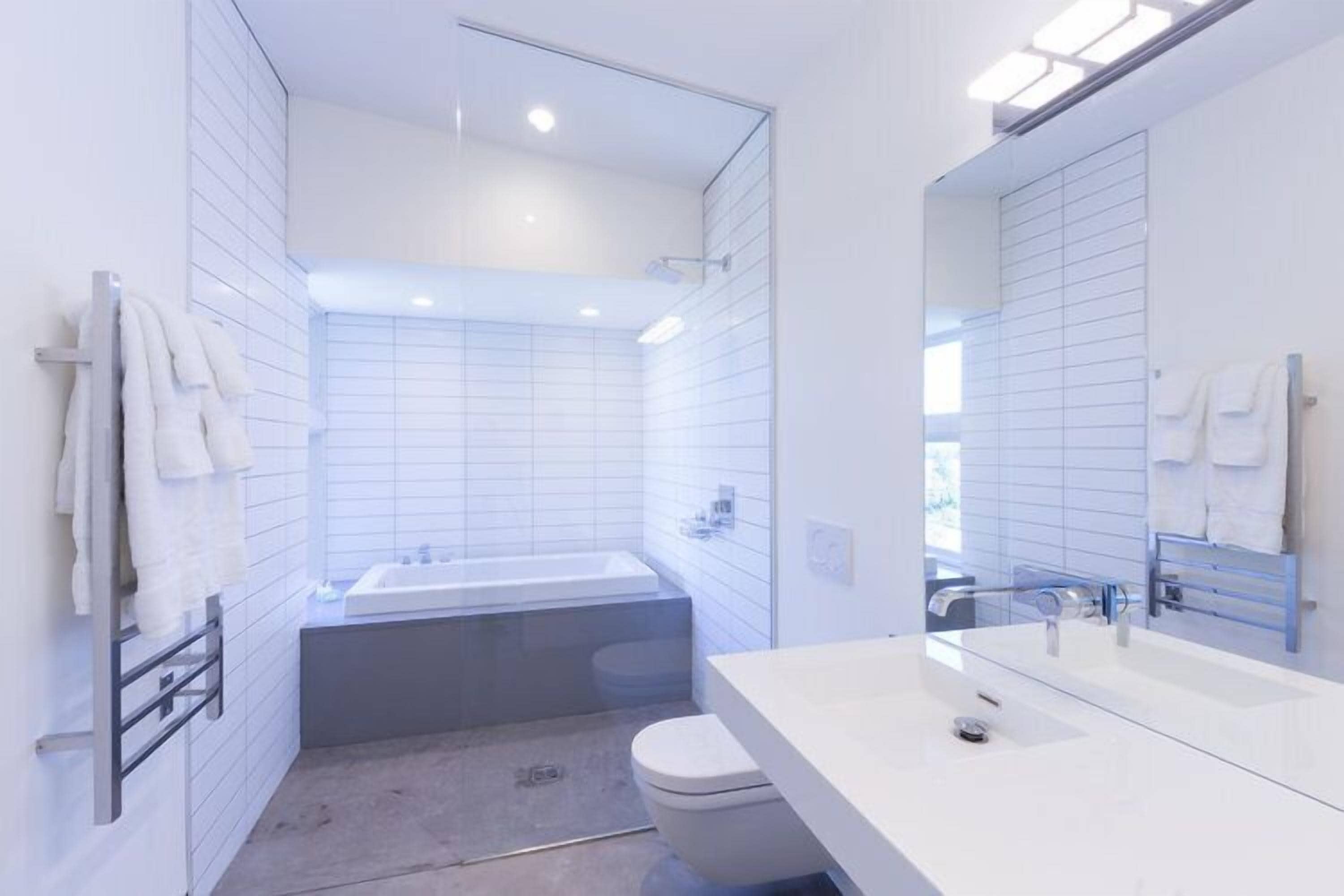 A modern bathroom with white tiles, a bathtub, a toilet, and a large mirror.